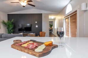Una sala de estar moderna con una tabla de madera que muestra diferentes carnes y quesos, una copa de vino y un televisor al fondo.