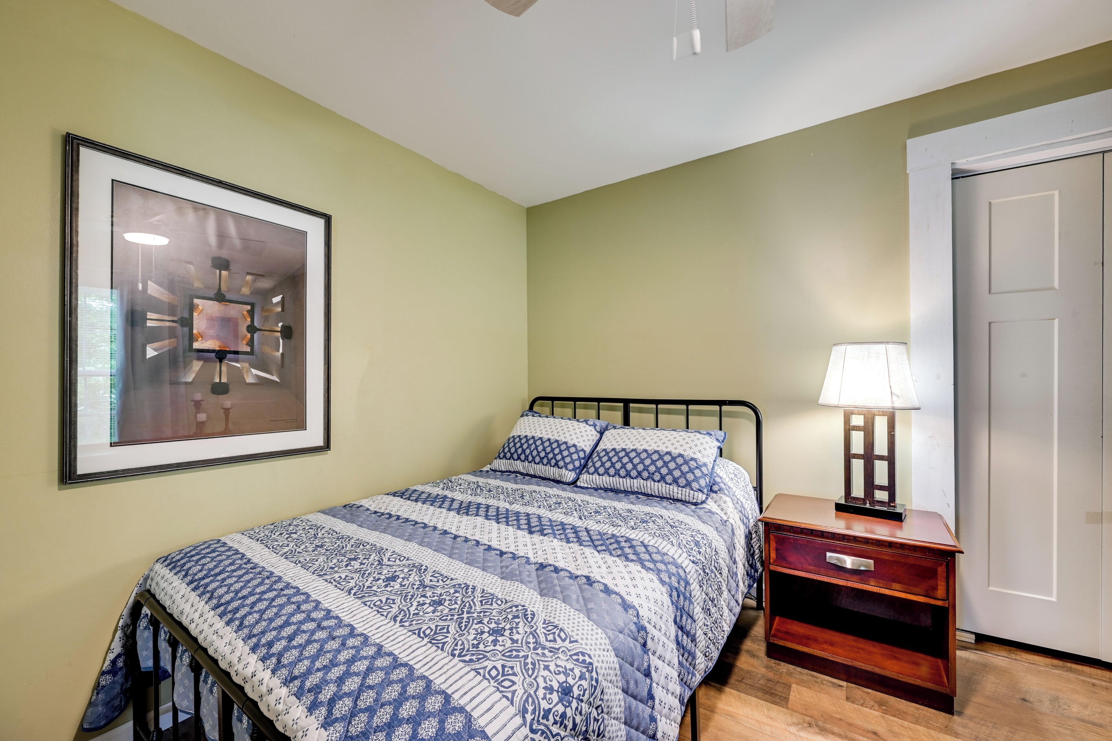 A bedroom with a bed, a nightstand, a lamp, a framed picture on the wall, and a door.