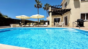 Ein Swimmingpool mit Liegestühlen und Sonnenschirmen, ein Grill und Palmen.