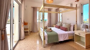 Ein Bett mit Baldachin, mit grüner Decke, ein Nachttisch mit Lampe und Blick auf das Meer durch das Fenster.