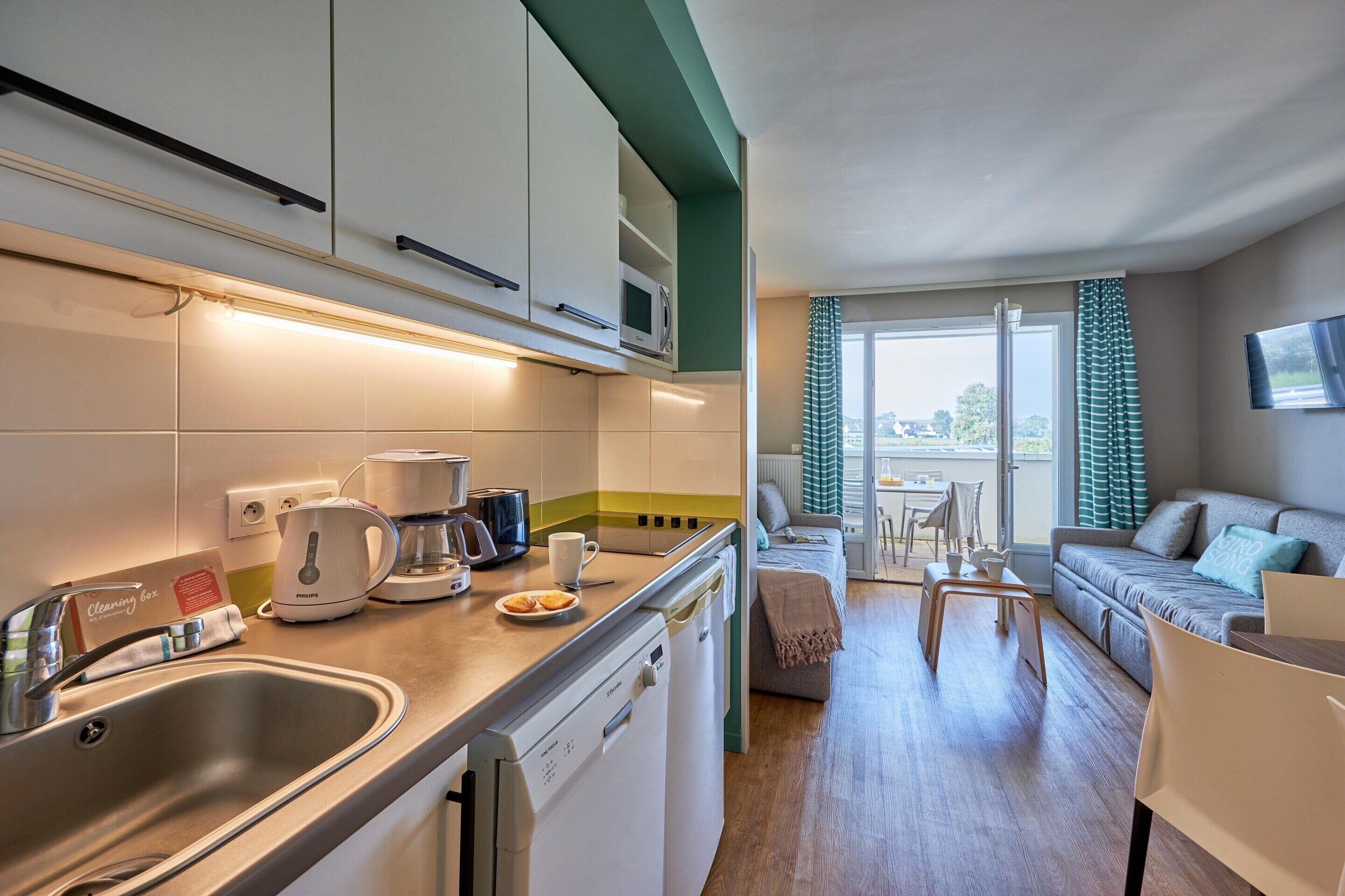 A modern kitchen with a dishwasher, a coffee maker, and a kettle. There is a dining area with a table and chairs, and a sofa in the background.