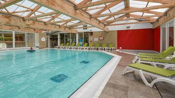 An indoor swimming pool with a glass ceiling, green lounge chairs, and a red wall.