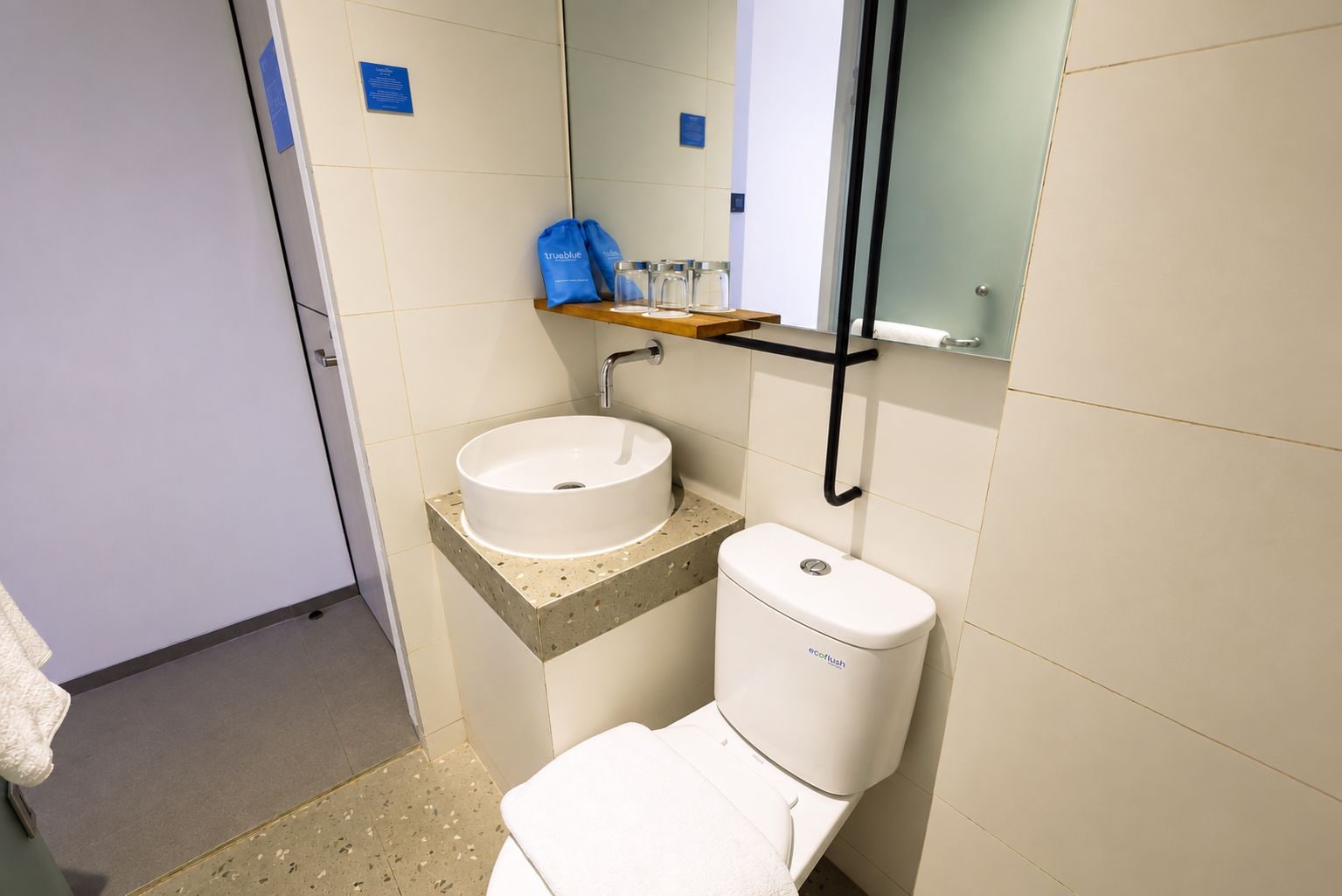 Ein Badezimmer mit einem weißen Waschbecken, einer Toilette und einem Spiegel. Über dem Waschbecken befindet sich ein Regal mit einer blauen Tasche und Gläsern.