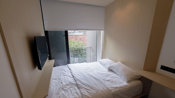 Ein Schlafzimmer mit Bett, einem an der Wand montierten Fernseher, einem Fenster mit Blick auf ein Gebäude und einem Balkon.