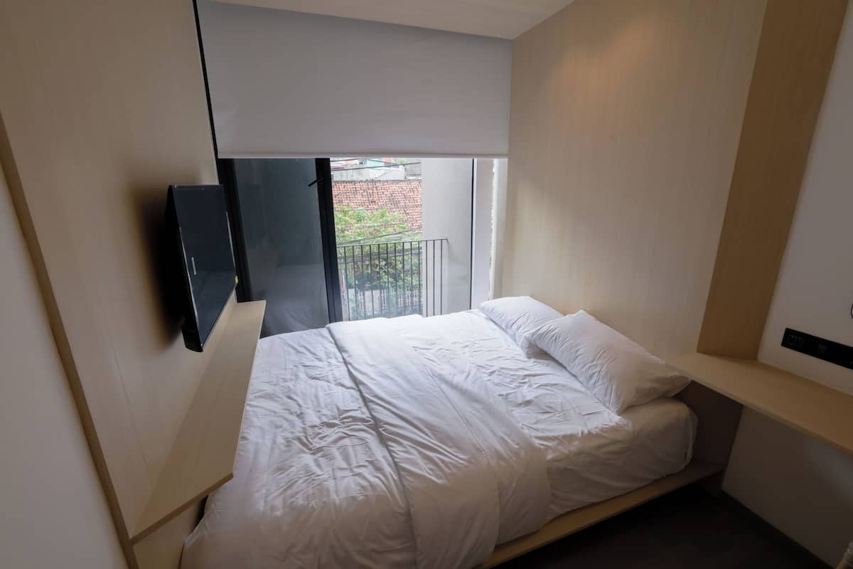 Un dormitorio con cama, televisor montado en la pared, ventana con vista a un edificio y balcón.