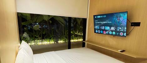 A modern hotel room with a bed, a mounted television displaying a movie, and a window with a view of greenery.