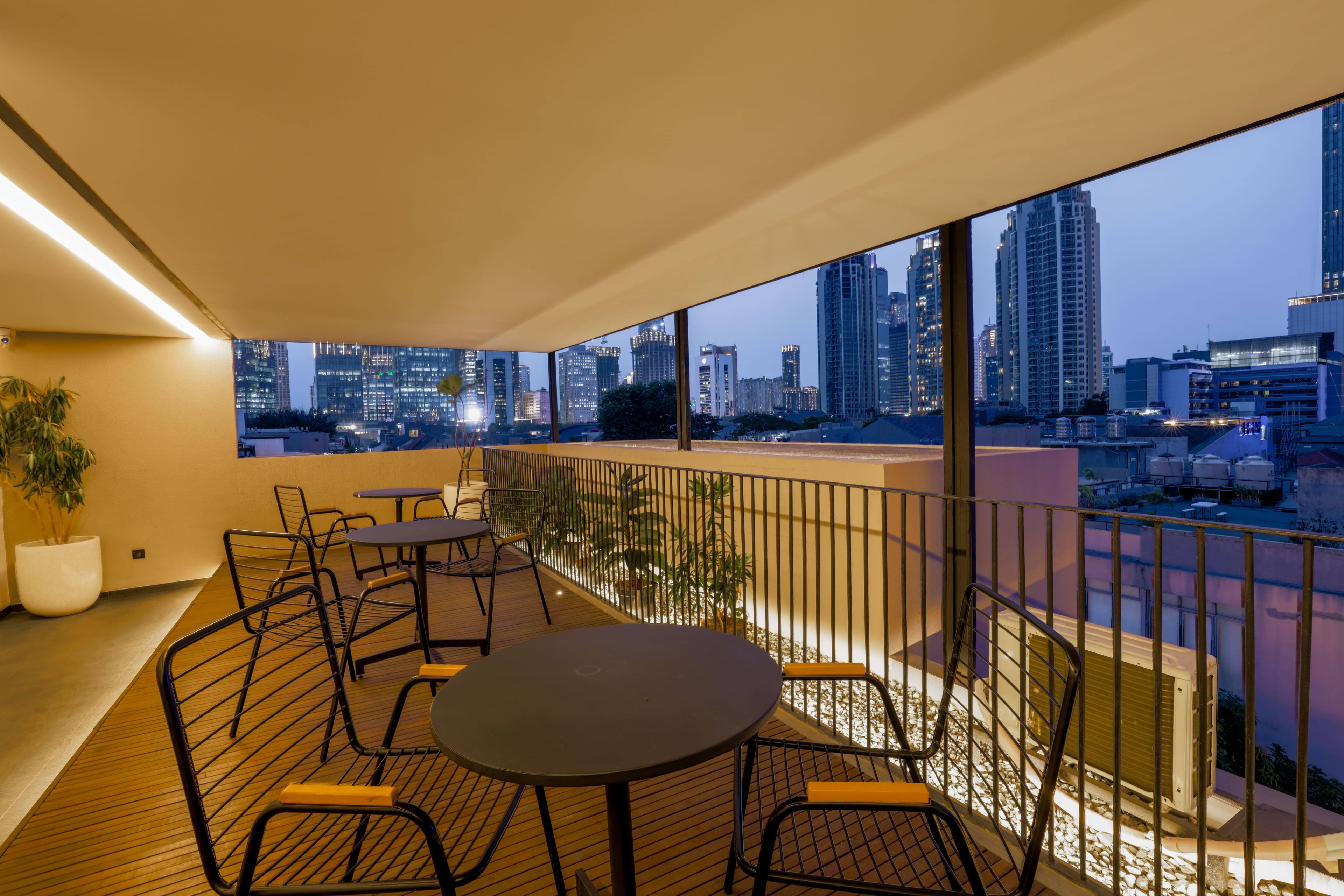 Een balkon in de schemering met uitzicht op de stad, met een tafel, stoelen, een bloempot en een hekwerk.