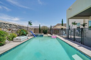 A swimming pool with a slide, surrounded by a stone wall, palm trees, and a modern building. - EP Oasis w/ Pool & Spa, Near I-10 & Hospitals (El Paso)
