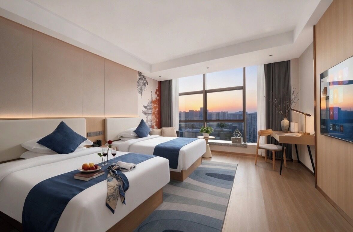 A hotel room with two beds, a desk, a chair, a TV, and a large window with a city view.
