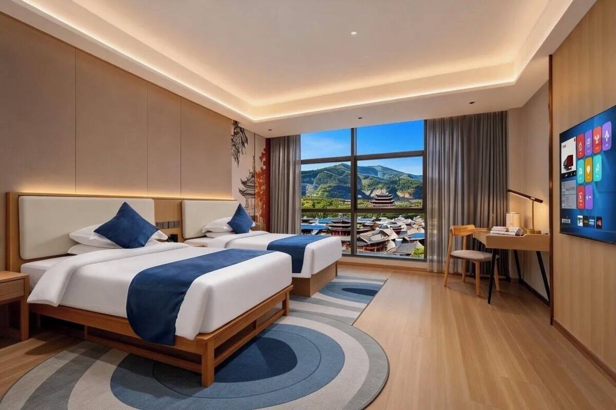 Habitación de hotel con dos camas, un escritorio, una silla, un televisor y un ventanal con vistas a un paisaje montañoso.