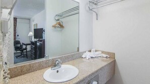 Une salle de bain d’hôtel avec un comptoir en granit, un lavabo blanc, un miroir, un porte-serviettes et un téléviseur en arrière-plan.