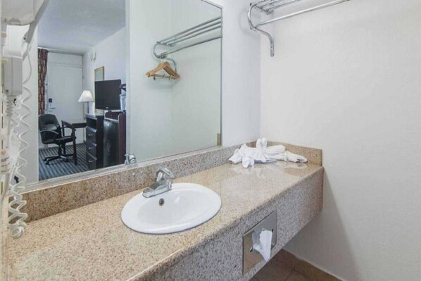 Un baño de hotel con encimera de granito, un lavabo blanco, un espejo, un perchero y un televisor al fondo.