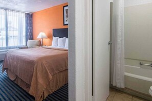A hotel room with a bed, a lamp, a window with curtains, and a bathroom visible through an open door.