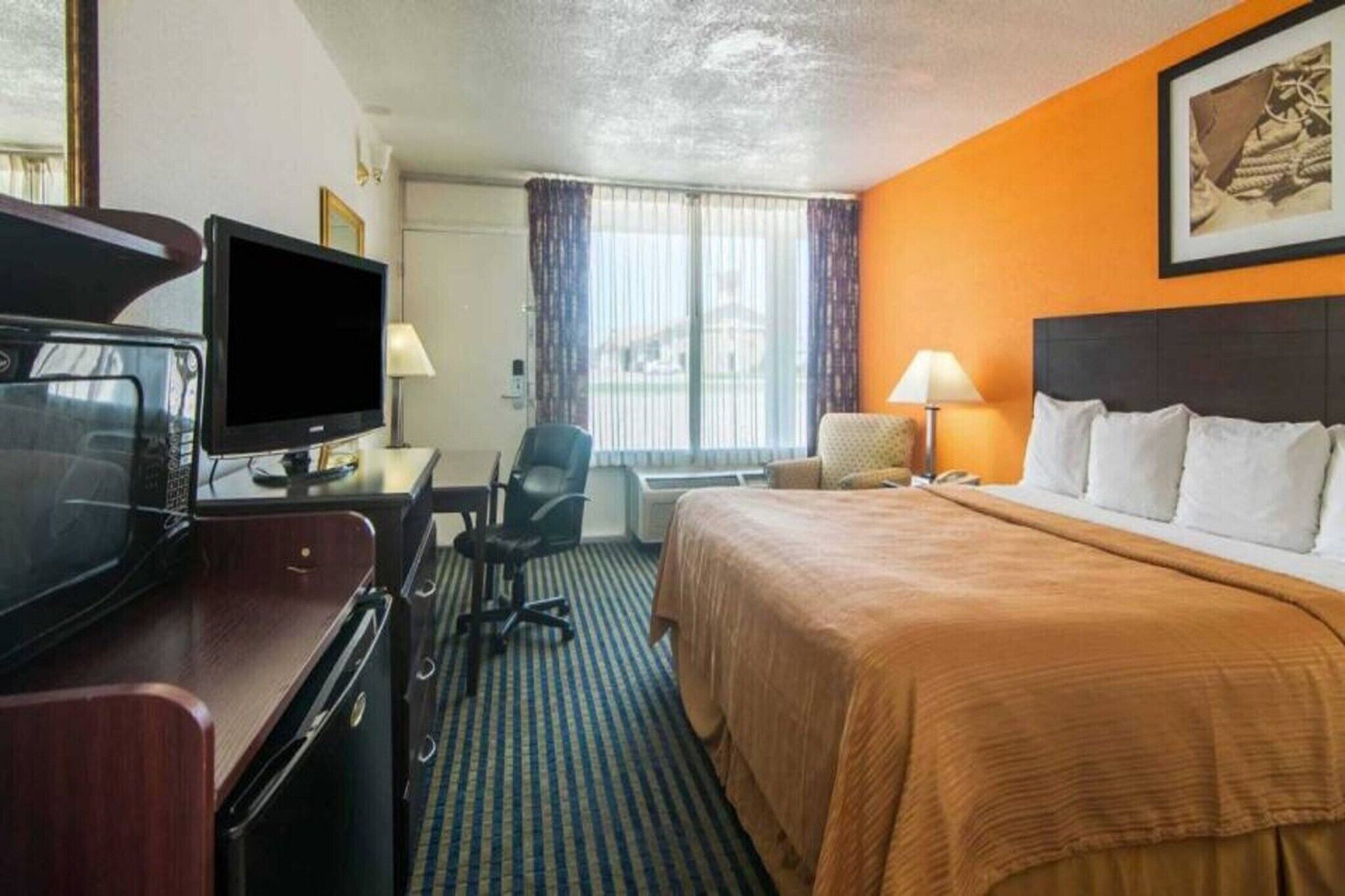Une chambre d’hôtel avec un lit, un bureau avec une télévision, une chaise, une lampe et une fenêtre avec des rideaux.