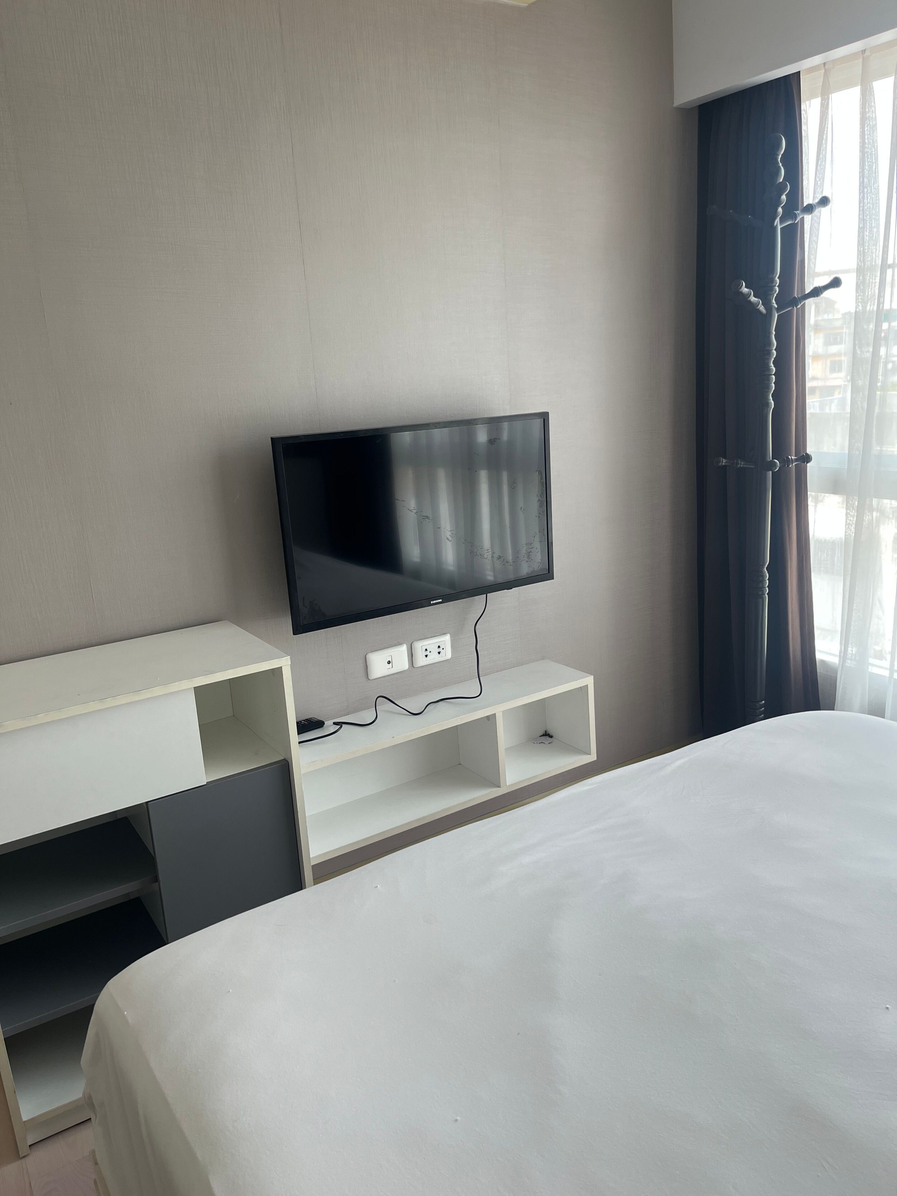 Habitación de hotel con una cama, un televisor sobre un pedestal blanco, una ventana con cortinas y un enchufe en la pared.