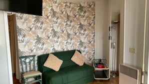 Et værelse med en grøn sofa, en stol, et glasbord, en radiator og en væg med et tropisk bladmotiv.