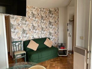 Ein Zimmer mit einem grünen Sofa, einem Sessel, einem Glastisch, einem Heizkörper und einer Wand mit tropischem Blattmuster.