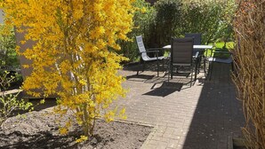 Een terras buiten met een tafel en stoelen, omringd door groen en een bloeiende gele boom.