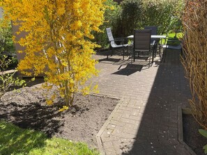 Ein möblierter Patio mit Tisch und Stühlen, umgeben von Grünpflanzen und einem blühenden gelben Baum.