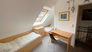 Een kleine, goed verlichte kamer met een schuine kap, een enkel bed, een bureau en een stoel.