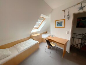 Ein kleiner, gut beleuchteter Raum mit schrägem Dach, einem Einzelbett, einem Schreibtisch und einem Stuhl.