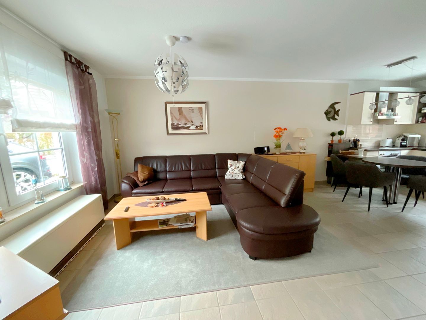 Ein Wohnzimmer mit braunem Ledersofa, einem Holztisch, einer Essecke mit Tisch und Stühlen, einer Küche mit modernen Geräten und einem großen Fenster mit Vorhängen.