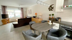 Een moderne woonkamer met een bank, een eettafel en stoelen, een keuken met kasten en apparatuur, en een televisie.