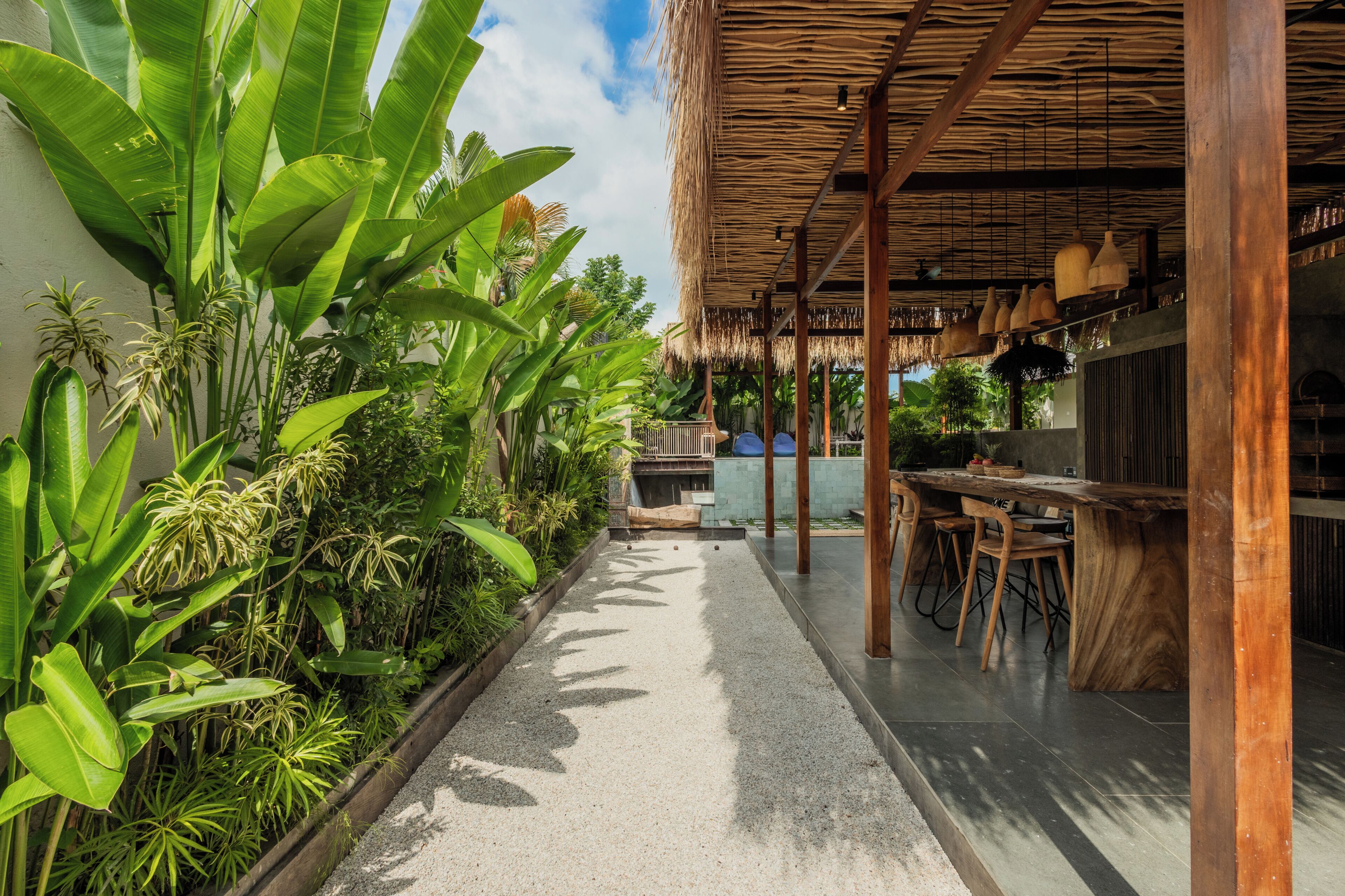 A covered outdoor area with a dining table and chairs, surrounded by lush greenery and a thatched roof structure.