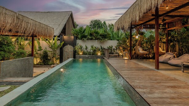 A pool with a wooden deck, thatched-roof structures, and lush greenery.