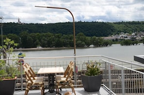 A balcony with a table and chairs, overlooking a river and a forested hill.