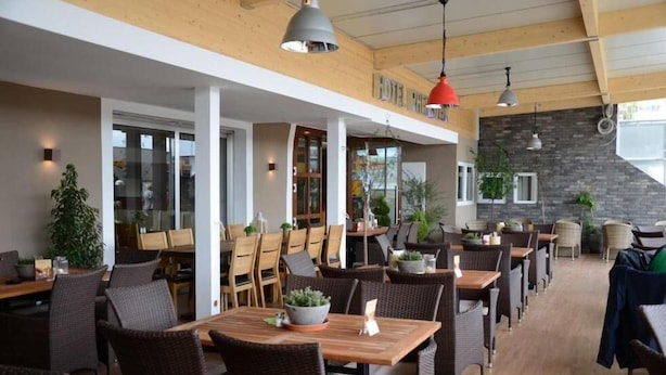 A spacious outdoor dining area with wooden tables and wicker chairs, large windows, and hanging pendant lights. - Hotel Rheinstein (Ruedesheim am Rhein)