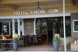 A hotel entrance with the name 'HOTEL RHENSTEIN' above the doors, outdoor seating with wicker furniture, and large potted plants.