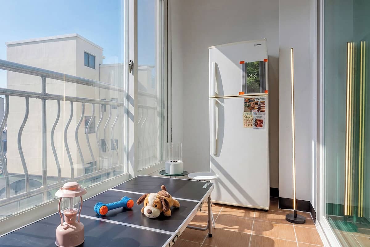 Un balcón con vista a edificios, una mesa con un perro de juguete y un farol, y un refrigerador con un letrero que dice 'Stay Alive'.