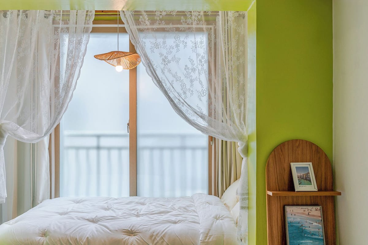 Un dormitorio con una cama, cortinas translúcidas, una lámpara colgante y un marco de foto sobre un estante de madera.