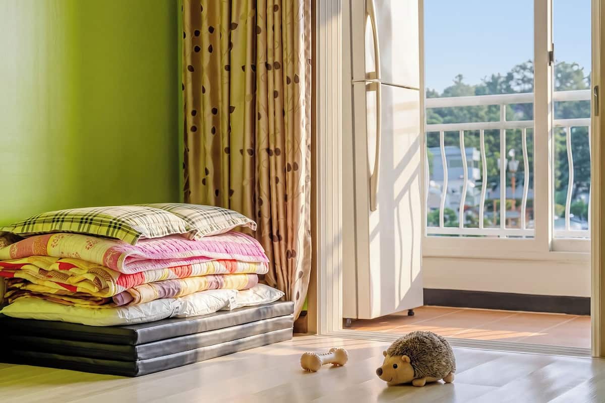 Una habitación con una cama, un montón de mantas dobladas, una ventana con vistas y un erizo de juguete en el suelo.