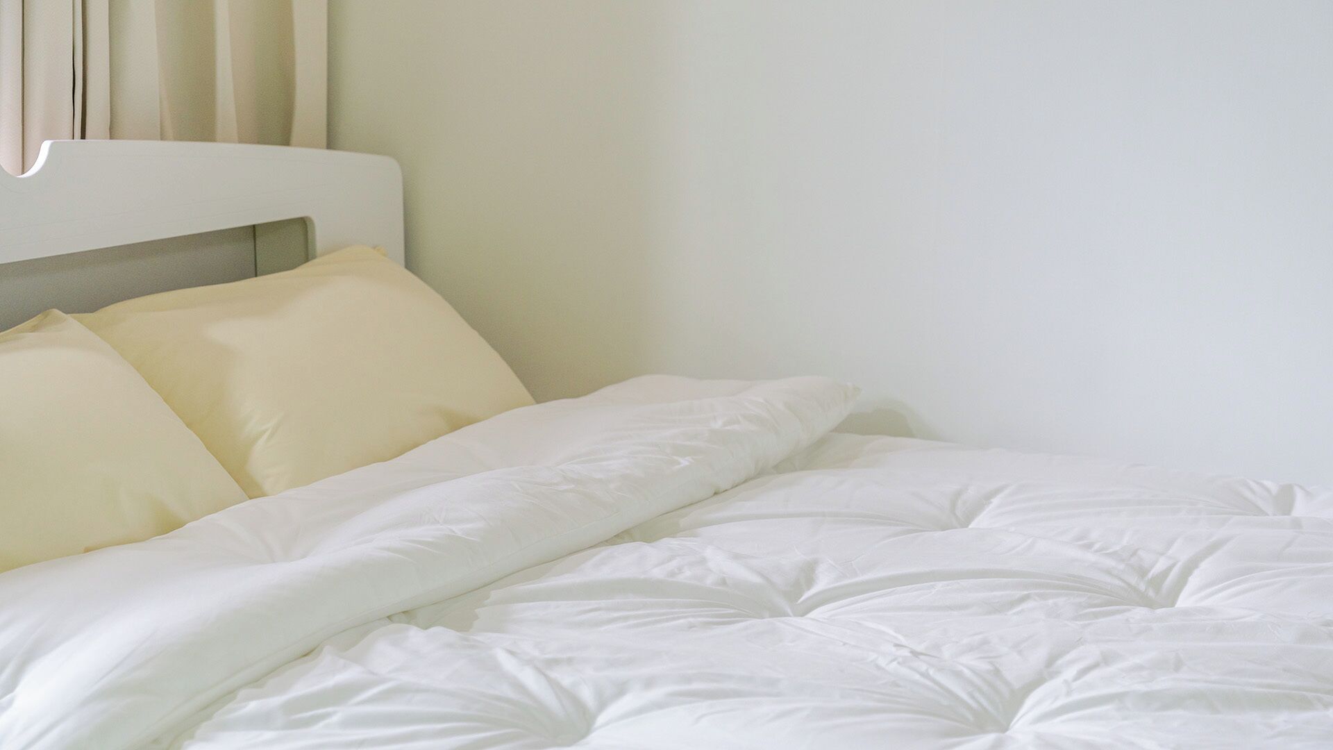 A neatly made bed with white bedding and pillows, a white headboard, and a plain wall.