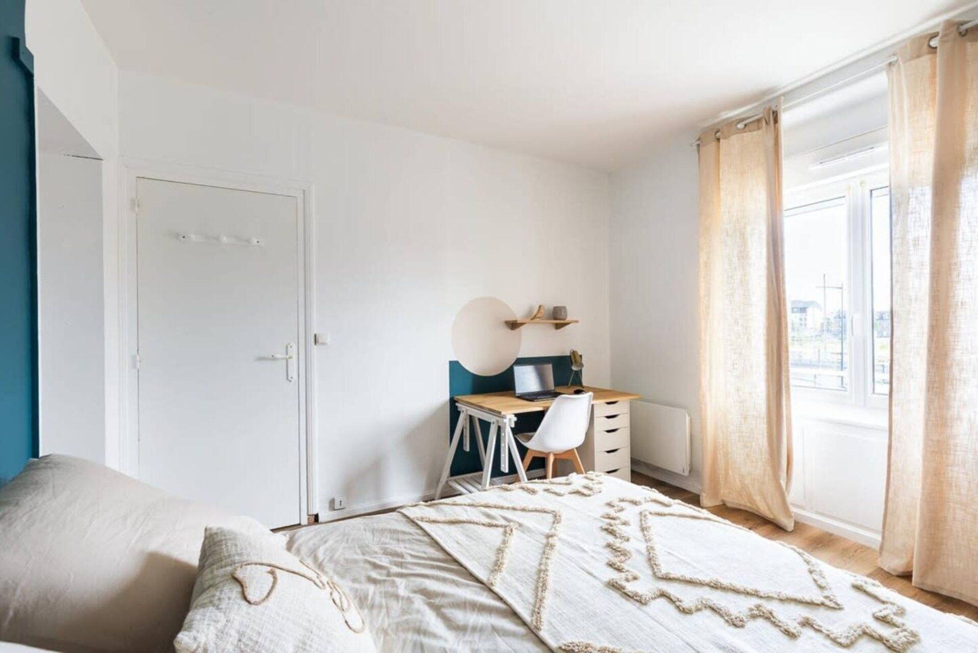 Une chambre à coucher avec un lit, un bureau et une chaise, une fenêtre avec des rideaux et une porte.