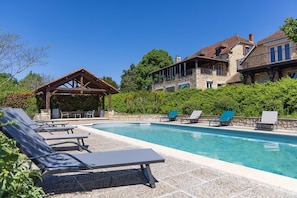 A swimming pool with lounge chairs, a wooden pergola, and stone buildings with balconies.