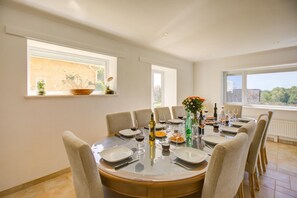 A dining room with a round table set for six, large windows, and a view of greenery outside.