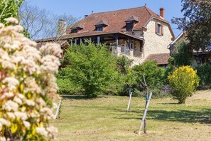 A stone house with a red-tiled roof, surrounded by greenery and a clear sky.
