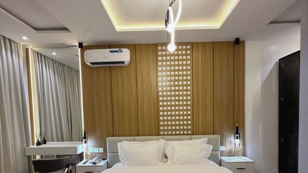 A modern hotel room with a large bed, a desk, a chair, a TV, and a wall-mounted air conditioner. - THE EVOLUTION HOTEL (Gombe)