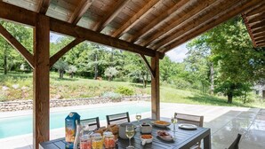 Un área de comedor al aire libre con vista a la piscina, pérgola de madera y una mesa dispuesta con comida y bebidas.