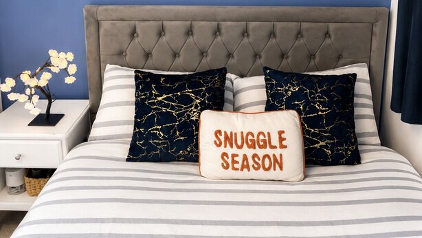 Ein Schlafzimmer mit einer gepolsterten Kopfteile, einem gerahmten Kunstwerk an der Wand und einem Kissen mit dem Aufdruck 'SNUGGLE SEASON'.