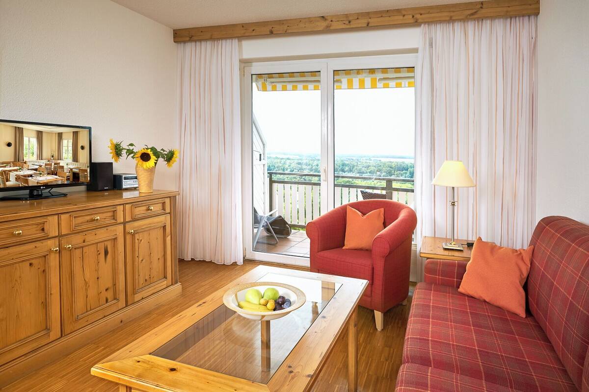 Una habitación con un armario de madera, un sillón rojo, un sofá, una mesa de centro con fruta y un balcón con vistas.