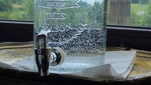 A clear glass jar labeled 'Hello SUMMER' with a metal tap, placed on a wooden surface with a scenic view of greenery and a structure in the background. - Les herbes folles de Oizy, bed and breakfast with terrace in the Ardennes. (Oizy)