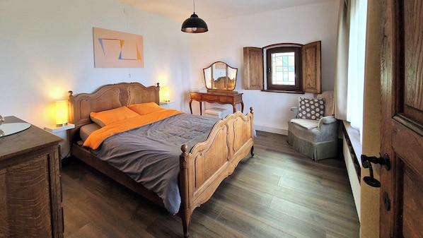 A bedroom with a wooden bed, bedside lamps, a dresser, a chair, a window with shutters, and a mirror on the wall.