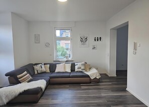 A modern living room with a sectional sofa, a white fluffy throw, and a view of the outside through a window. - Holiday Home 'Ritah' with Shared Terrace, Private Garden and Wi-Fi (Idar-Oberstein)