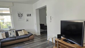 Moderni olohuone, jossa on harmaa sohva, litteänäyttöinen televisio puisen jalustan päällä ja taustalla ruokailutila.