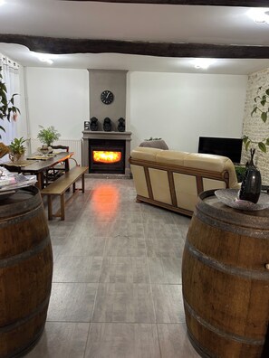 Ein Wohnzimmer mit Kamin, einer Couch, Holzfässern, einem Esstisch mit Stühlen und einer Wanduhr.