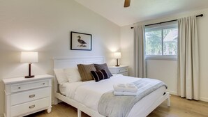 Una cama bien hecha con almohadas, una mesita de noche con lámpara, un cuadro en la pared y una ventana con cortinas.